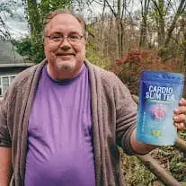 A smiling middle-aged man holding a pouch of Cardio Slim Tea outdoors, showcasing a customer testimonial for heart health and weight management